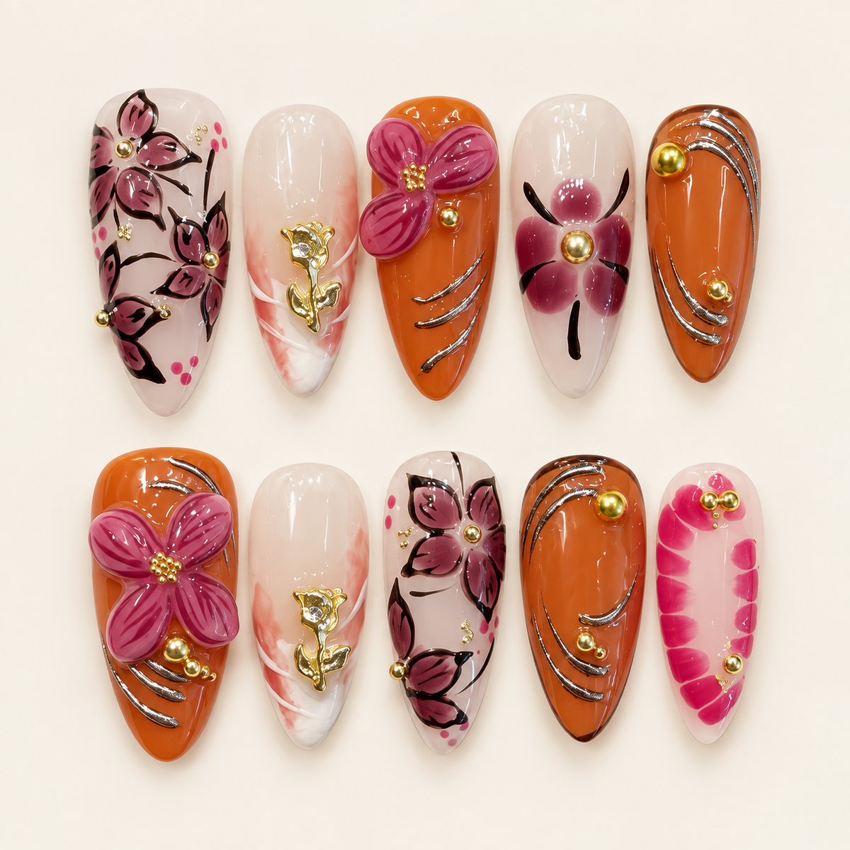 Dix press on nails amande orange et nude, décorés de fleurs framboise, de roses dorées et de courbes argentées, sur fond ivoire.