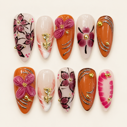 Dix press on nails amande orange et nude, décorés de fleurs framboise, de roses dorées et de courbes argentées, sur fond ivoire.