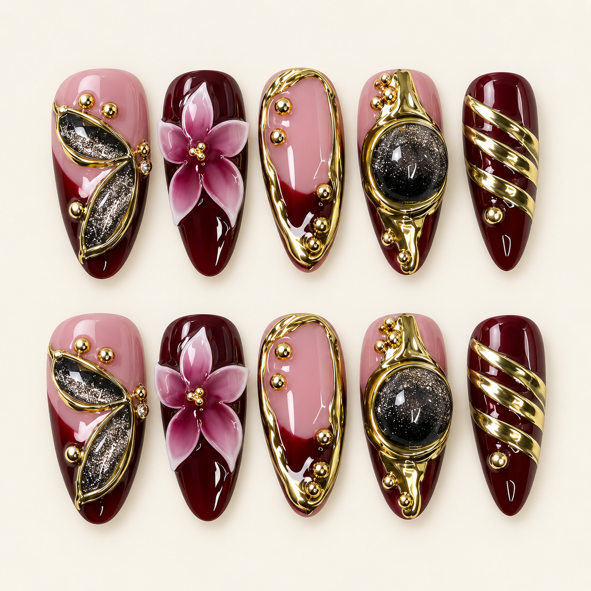 Dix press on nails amande bordeaux et rose poudré, décorés de fleurs 3D, de contours dorés et d’accents noirs scintillants, sur fond ivoire.