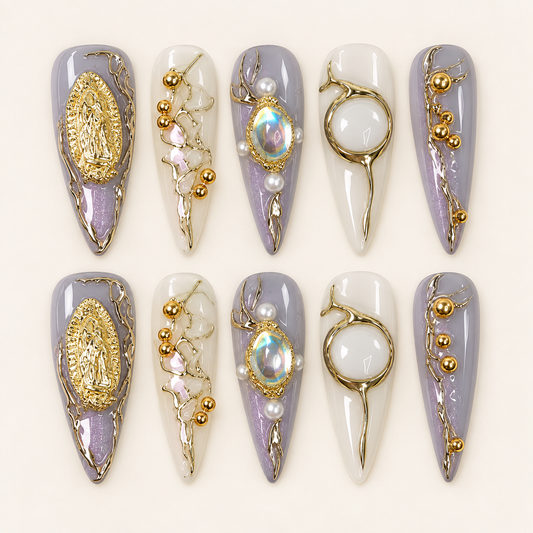 Dix press on nails longs et pointus lilas et blanc laiteux, décorés de médaillons dorés, de cabochons irisés et de perles, sur fond ivoire.
